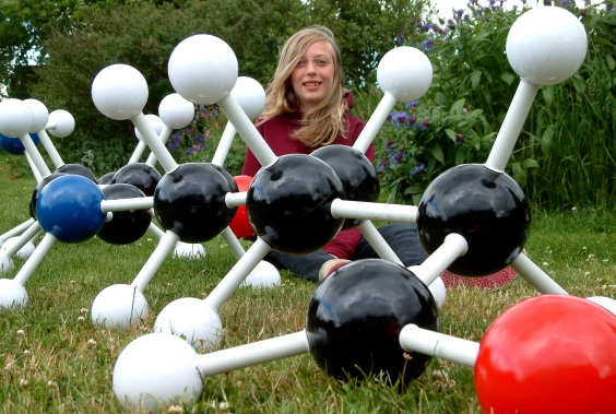 A girl sitting on grass behind a giant molecular model of nylon-6 1